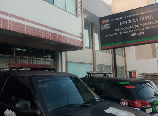 fachada e placa da delegacia, com duas viaturas estacionadas na frente