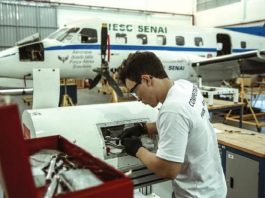 rapaz utilizando óculos e luvas mexe em turbina de aeronave em hangar; há um avião parado ao fundo
