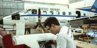 rapaz utilizando óculos e luvas mexe em turbina de aeronave em hangar; há um avião parado ao fundo