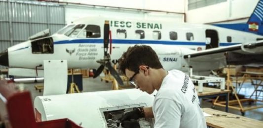 rapaz utilizando óculos e luvas mexe em turbina de aeronave em hangar; há um avião parado ao fundo