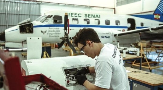 rapaz utilizando óculos e luvas mexe em turbina de aeronave em hangar; há um avião parado ao fundo