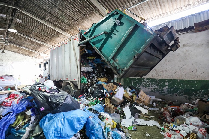 caminhão de lixo abre a parte traseira despejando grande quantidade de material reciclável em galpão