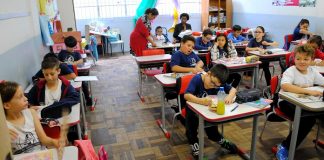 crianças sentadas em carteiras em sala de aula e duas professoras ao fundo