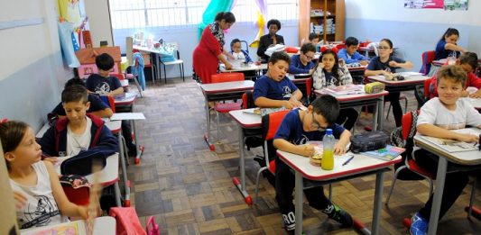 crianças sentadas em carteiras em sala de aula e duas professoras ao fundo