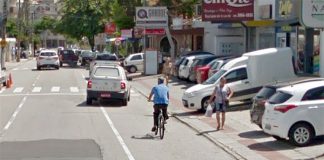 ciclista anda sobre uma via de três pistas em bairro comercial; há carros estacionados ao lado