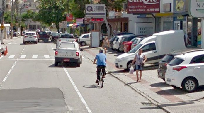 ciclista anda sobre uma via de três pistas em bairro comercial; há carros estacionados ao lado