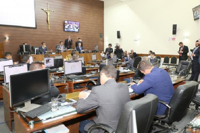 vereadores sentados em suas mesas com computadores em sessão da câmara; alguns de pé ao fundo