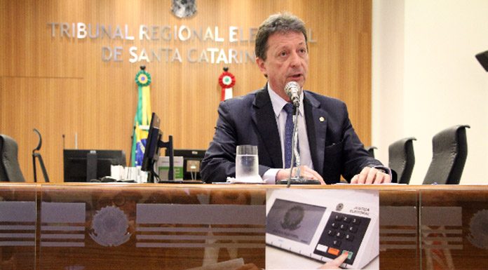 desembargador fala ao microfone numa bancada do tribunal, com um pequeno cartaz da urna eletrônica à frente