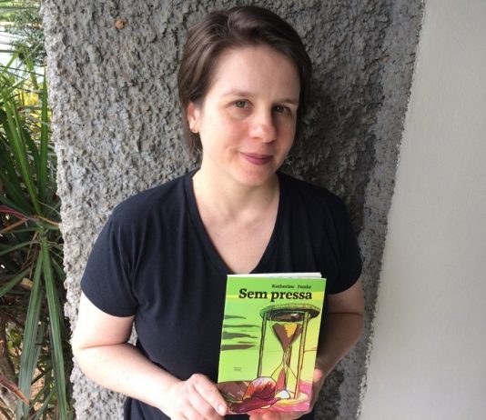 escritora katherine olha para a câmera posando com o livro em mãos e apoiada em uma parede