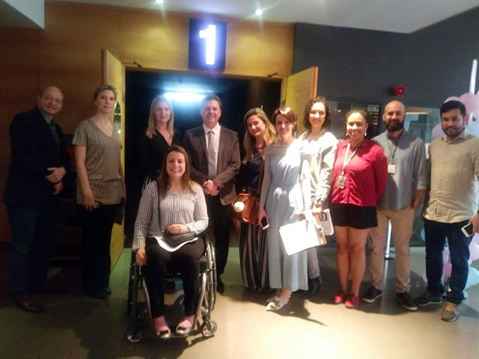 grupo de homens e mulheres posa para foto na porta de uma sala de cinema.; há uma mulher à frente em cadeira de rodas