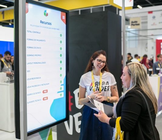 duas mulheres conversam entre si à frente de um grande tela em uma feira