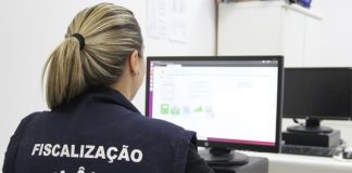 mulher com colete de fiscalização da vigilância sanitária é vista de costas trabalhando em computador