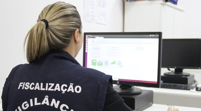 mulher com colete de fiscalização da vigilância sanitária é vista de costas trabalhando em computador