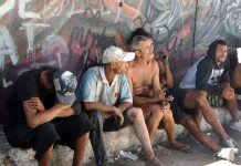 homens moradores de rua sentados em pequena sarjeta junto a muro grafitado olham na mesma direção