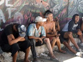 homens moradores de rua sentados em pequena sarjeta junto a muro grafitado olham na mesma direção