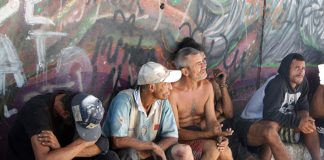 homens moradores de rua sentados em pequena sarjeta junto a muro grafitado olham na mesma direção
