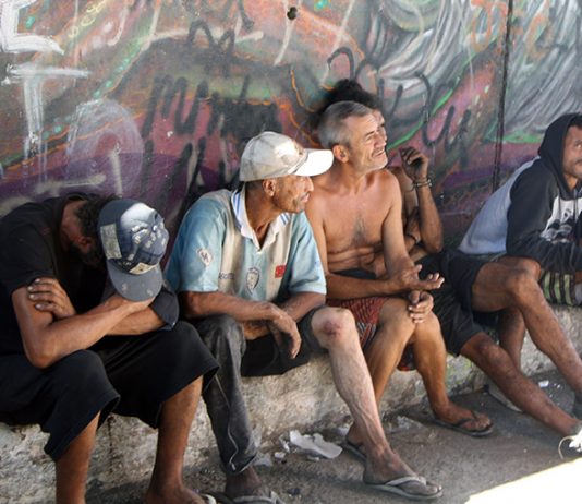 homens moradores de rua sentados em pequena sarjeta junto a muro grafitado olham na mesma direção