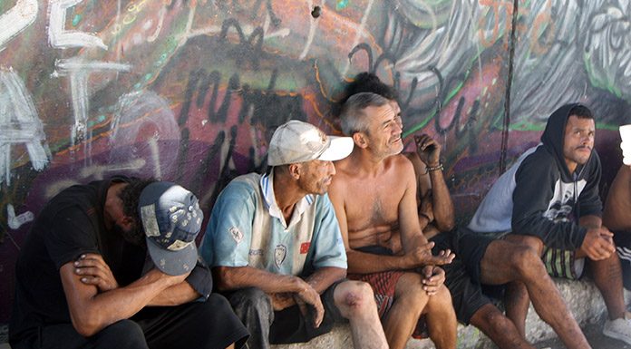 homens moradores de rua sentados em pequena sarjeta junto a muro grafitado olham na mesma direção