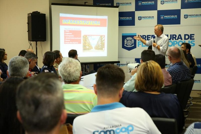 prefeito em pé gesticula com um microfone na mão ao lado de uma tela com dados e pessoas observam sentadas