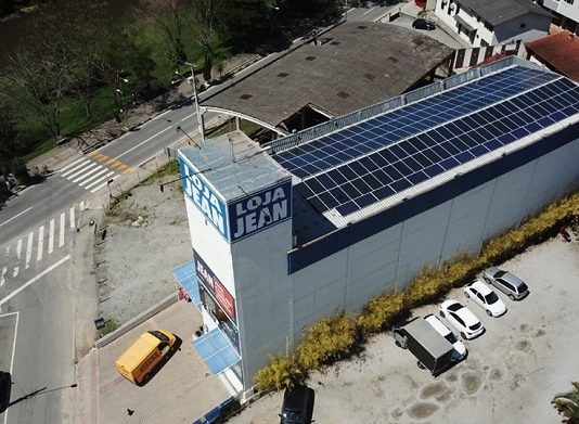 Energia solar ganha espaço em SC loja vista de cima, em foto de drone, mostrando todos os painéis solares no teto do prédio; há um logo "loja jean"