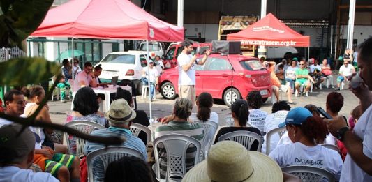 Comcap e Sintrasem fecham acordo coletivo de trabalho trabalhadores com camisas escritas "dasta-base" sentados assistem a um homem discursas; carro e tenda ao fundo do sindicato