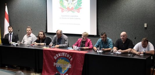 diversas pessoas em uma bancada à frente de um auditório; há uma bandeira do movimento de população de rua sobre a bancada; deputada carminatti ao centro fala no microfone