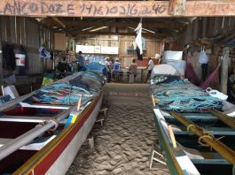 dois barcos guardados dentro de um rancho de pesca, com chão de areia; algumas pessoas ao fundo do rancho