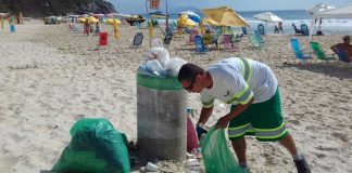 trabalhador da comcap se abaixa para pegar lixo com sacola na mão ao lado de lixeira em uma praia