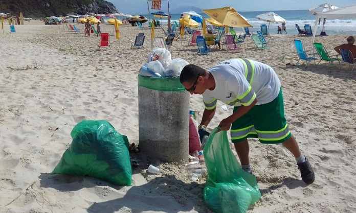 trabalhador da comcap se abaixa para pegar lixo com sacola na mão ao lado de lixeira em uma praia