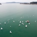 imagem aérrea do mar esverdeado com diversos barcos à vela espalhados; ao fundo algumas montanhas da ilha de sc