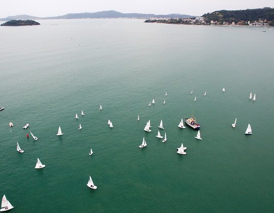 imagem aérrea do mar esverdeado com diversos barcos à vela espalhados; ao fundo algumas montanhas da ilha de sc