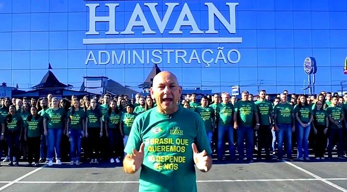 luciano hang fala para a câmera com funcionários uniformizados atrás em frente à uma loja da havan