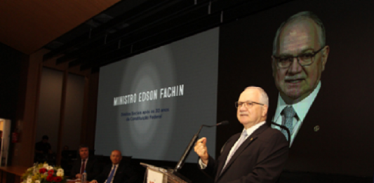 ministro fachin fala ao microfone em uma tribuna, ao lado de uma bancada e telão com seu nome