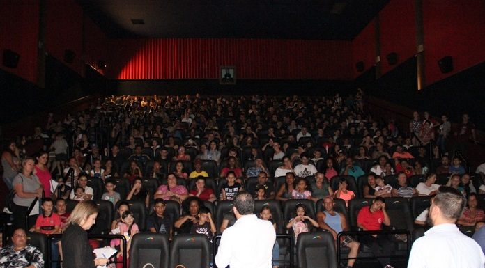 prefeito visto de costas ao centro da foto fala para um auditório lotado principalmente de crianças; mais algumas pessoas ao seu lado