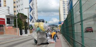 homem se abaixa para ajeitar grande quantidade de sacolas e materiais em um carrinho de coleta parado ao lado de calçada