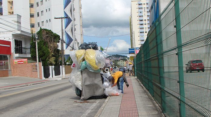 homem se abaixa para ajeitar grande quantidade de sacolas e materiais em um carrinho de coleta parado ao lado de calçada