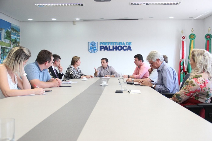 prefeitos reunidos em volta de uma grande mesa, com o prefeito camilo na ponta gesticulando e falando e atrás um logo da prefeitura de palhoça