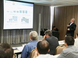 cosme fala ao microfone olhando para um telão em frente a uma plateria masculina sentada que observa em salão cheio de cortinas