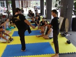 dois homens mostram golpes em um tatame onde uma turma de mulheres está sentada assistindo