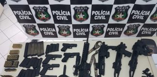 mesa cheia de armas, metralhadoras e pentes de munição à frente de um painel com logos da polícia civil