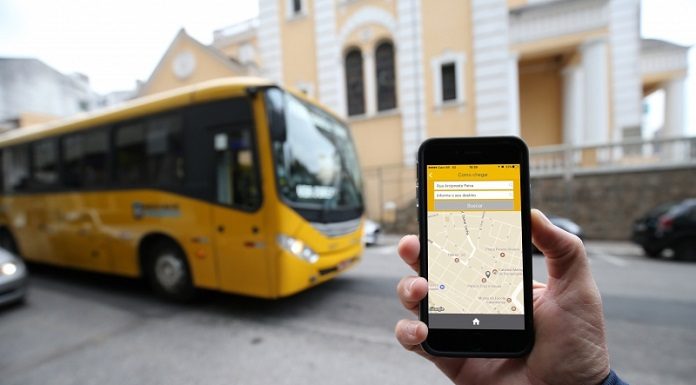 mão em frente à câmera segurando celular com aplicativo aberto e um ônibus amarelo passando atrás