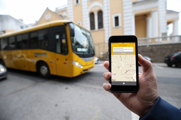 mão em frente à câmera segurando celular com aplicativo aberto e um ônibus amarelo passando atrás