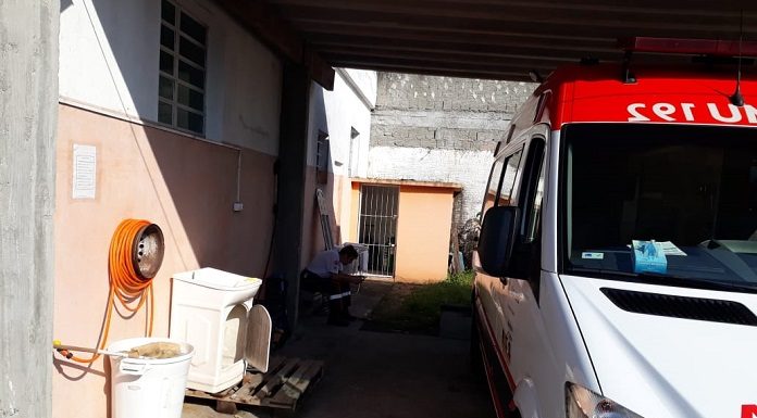 ambulância estacionada em garagem precária junto à uma casa, com máquina de lavar roupa antiga e outros objetos; há um homem sentado ao fundo na sombra