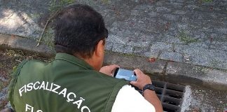 fiscal da floram, usando colete, se abaixa para tirar foto com celular de um bueiro