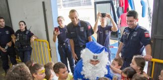 papai noel na cor azul agachado e rodeado de crianças observado por agentes sorridentes da guarda municipal