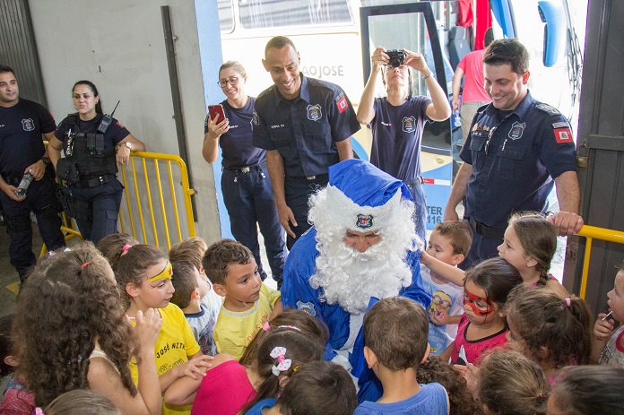 papai noel na cor azul agachado e rodeado de crianças observado por agentes sorridentes da guarda municipal