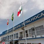 fachada do prédio atual da prefeitura de palhoça com as bandeira de sc, br e do município à frente