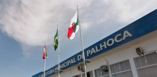 fachada do prédio atual da prefeitura de palhoça com as bandeira de sc, br e do município à frente