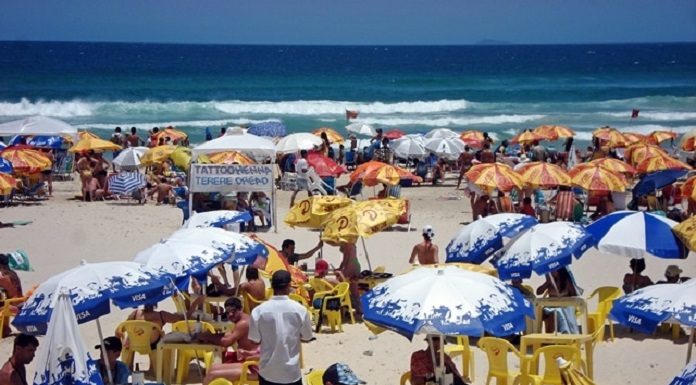 dezenas de mesas e cadeiras com guarda-sóis espalhados por um praia e pessoas circulando