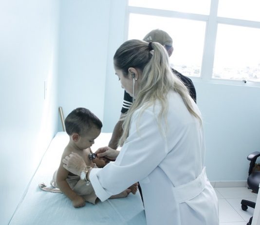 médica escuta o peito de uma criança sentada na maca do consultório com um parente ao lado